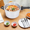 A white and silver 16-piece pressure cooker with black handles sits on a wooden dining table, filled with a colorful noodle soup. Surrounding the cooker are small bowls of ingredients, a folded white cloth, and a white plate with silver cutlery.