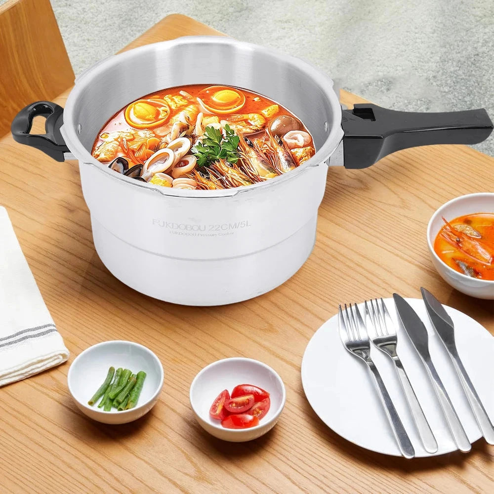 A white and silver 16-piece pressure cooker with black handles sits on a wooden dining table, filled with a colorful noodle soup. Surrounding the cooker are small bowls of ingredients, a folded white cloth, and a white plate with silver cutlery.
