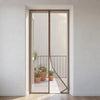 A modern magnetic door screen with a thin brown metal frame and vertical slats, shown installed in a doorway leading to a sunlit balcony with potted plants