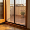 A close-up view of a sliding glass door fitted with a fine mesh screen, set in a warm indoor environment with wooden flooring and a sunlit outdoor patio visible behind it.