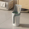 A white and gray automatic sifting trash can with a green-lined inner bin, shown on a tiled floor next to a wooden cabinet.