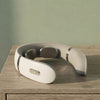 A neck and shoulder massager in a warm off-white color, resting on a wooden surface against a textured sage green wall.