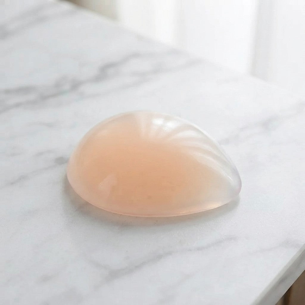A translucent peach-colored silicone breast implant or gel pad resting on a white marble surface.