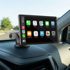 A car stereo touchscreen display mounted on a dashboard with a suction cup, showing a home screen with app icons. The device is positioned inside a car with the windshield and road visible in the background.
