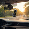 A black car windshield suction mount with an adjustable camera holder, shown installed on the rearview mirror of a car with a road and trees visible through the windshield.