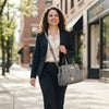 A woman with curly brown hair walks down a city street wearing a navy blazer, cream blouse, and dark trousers, carrying a structured gray handbag with gold-tone hardware.