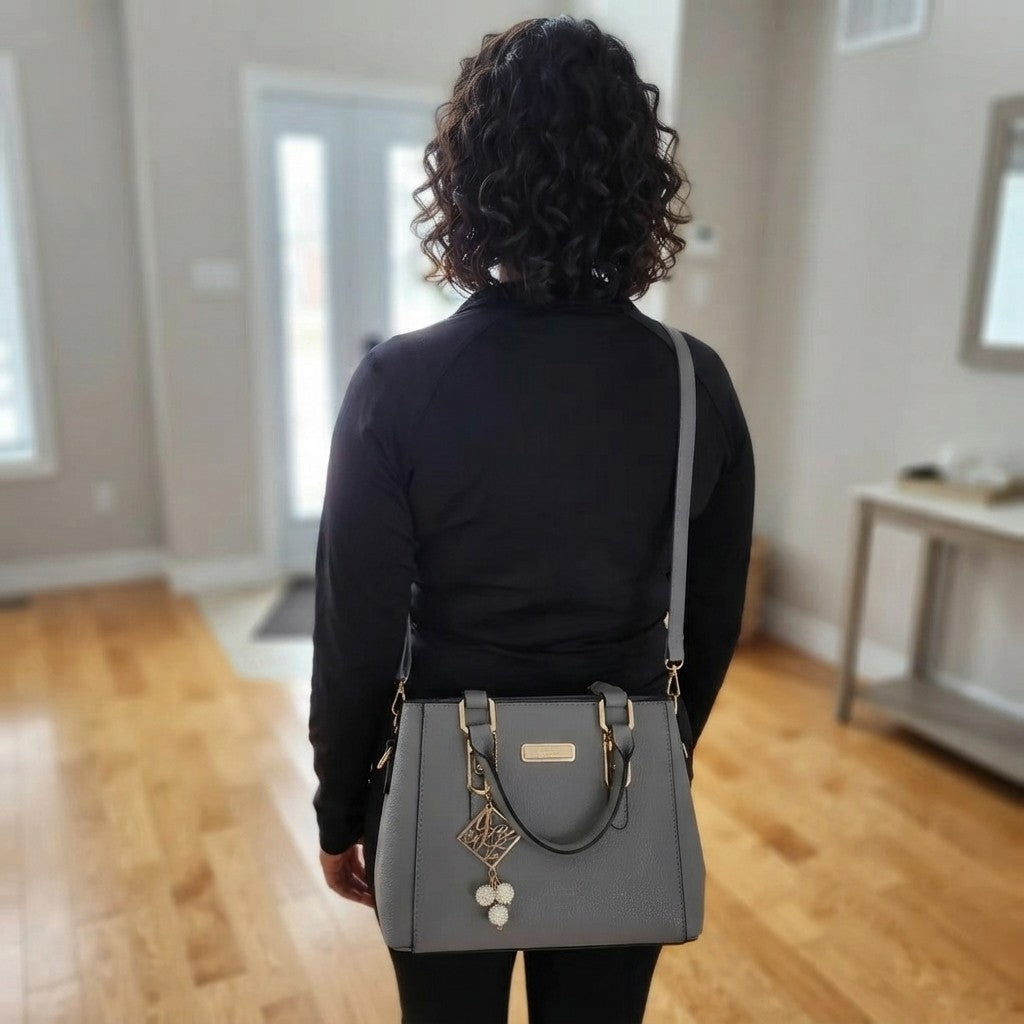 A woman viewed from behind wearing a black long-sleeve top and black pants, carrying a gray shoulder bag with gold-tone hardware and a hanging charm in a bright room with wood floors.