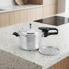 A stainless steel pressure cooker with black handles sits on a speckled kitchen countertop, with a matching metal steamer insert placed beside it. A blurred kitchen interior is visible in the background.