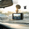 A black car dash cam with a suction cup mount and a small rear camera accessory, shown installed on a car windshield with a blurred road scene behind it.