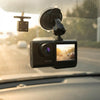 A black 1080p dash camera with a built-in screen, mounted on a black suction cup bracket attached to a car windshield. The camera is shown in a real-world car environment with a blurred road and warm sunset light filtering through the windshield.