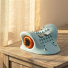 A light blue ergonomic desk accessory with an orange circular detail and silver buttons, resting on a wooden table next to a beige curtain.